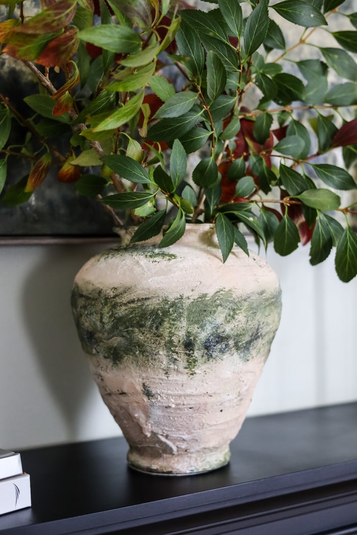 rustic looking vase tan and green filled with green branches on a mantle.