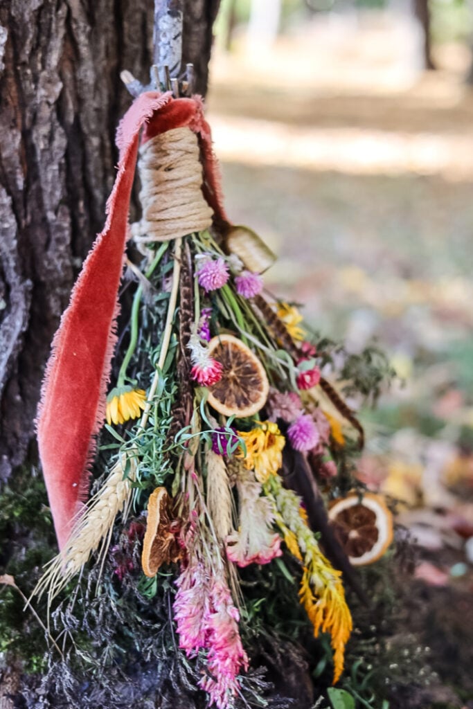 a witch broom made with flowers, greenery, herbs, and orange slices leaning against a tree